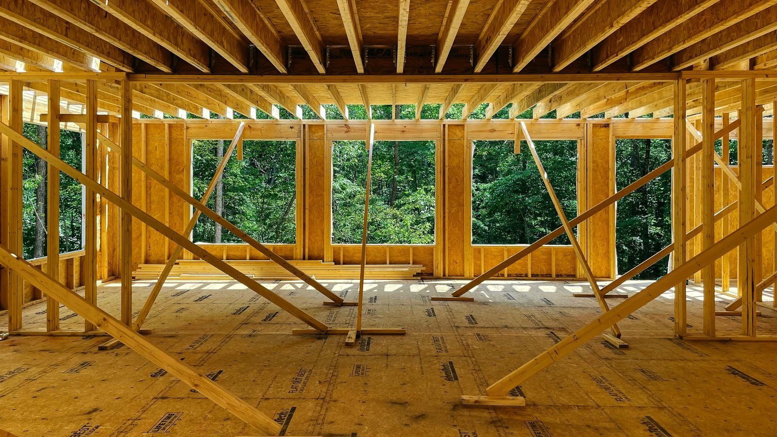 New construction home during framing — exposed wood studs and joists, multiple window openings facing a forest.