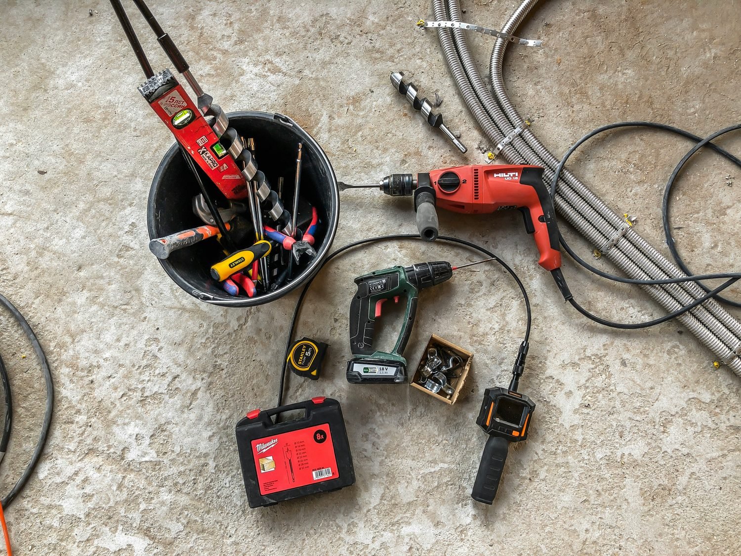 Top-down view of electrical and plumbing tools on a concrete floor — a rotary hammer, cordless drill, tape measure, inspection scope, and flexible conduit laid out beside a bucket of bits.
