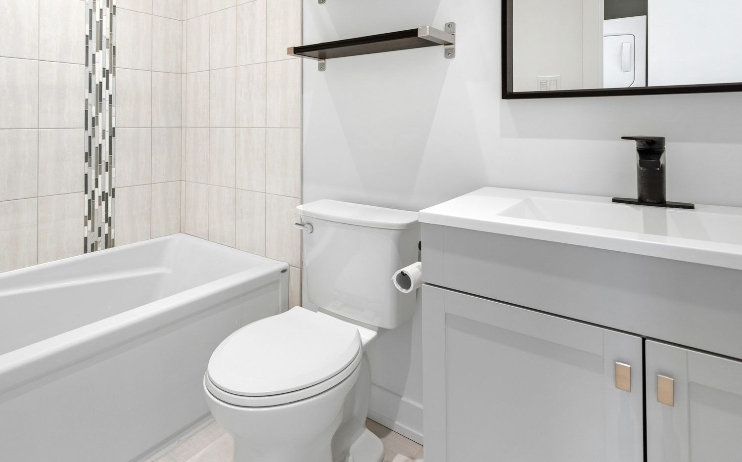 Remodeled bathroom — white tile tub surround, light gray vanity with quartz countertop and black faucet, and a black-framed mirror above the sink.