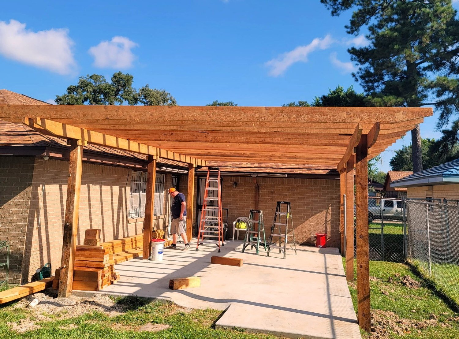 Patio cover under construction: cedar post-and-beam frame with rafters going up over a poured concrete slab on the back of a brick home